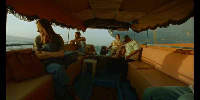 Ein überdachtes Boot auf dem Wasser, vorn sitzt eine Frau, im Hintergrund drei Männer. Ganz vorn sieht man die Beine einer weiteren Person.