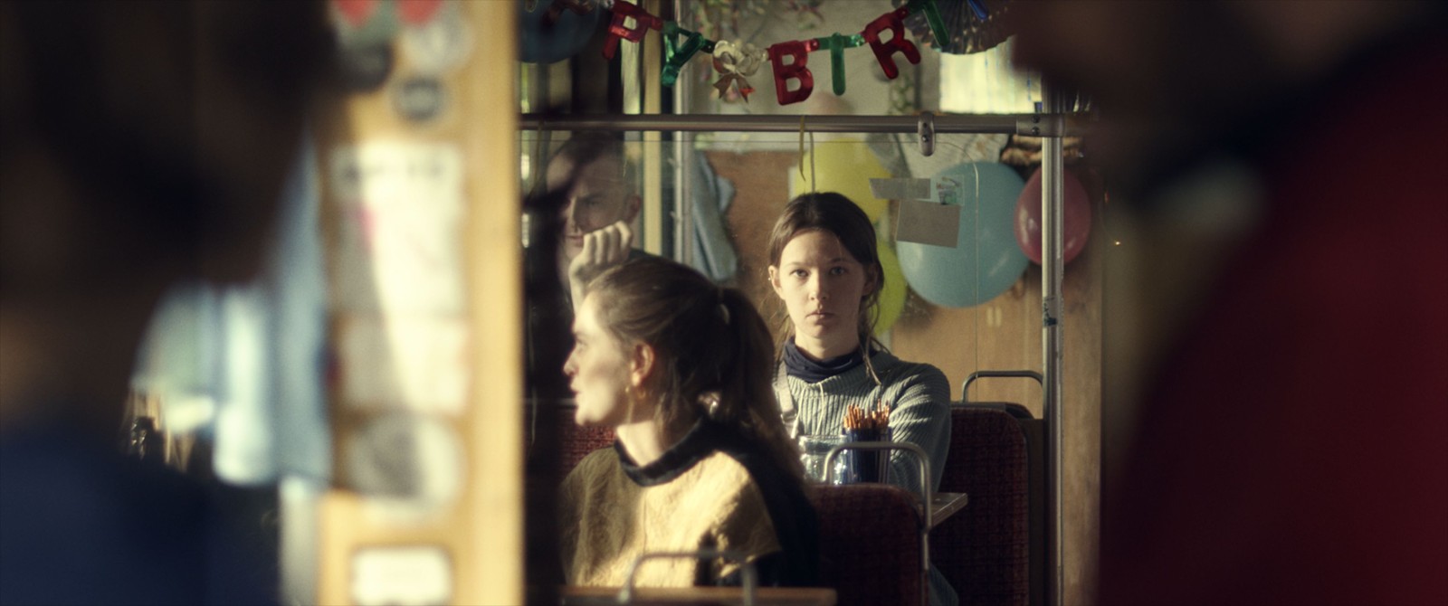 Zwei junge Frauen sitzen hintereinander. Die hintere schaut direkt in die Kamera, über ihr eine bunte Buchstabenkette mit "Happy Birthday".