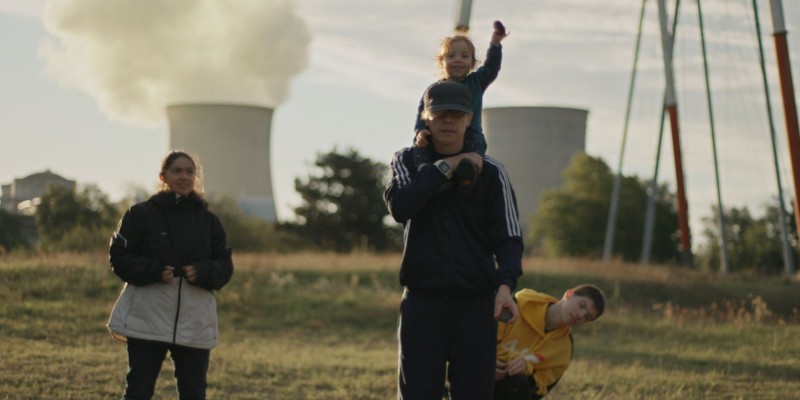 Ein Mann mit einem Kind auf der Schulter, ein weiteres sitzt hinter ihm und schaut an ihm vorbei, neben ihm steht eine Frau. Im Hintergrund ist ein Atomkraftwerk.
